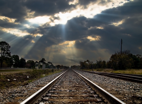 A train track leads into the distance with a dramatic dark sky with sunlight bursting through