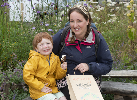 A woman and child sit in a wildflower garden smiling, the woman holds a wildchild paper bag and wears a head scarf, the child holds an icecream