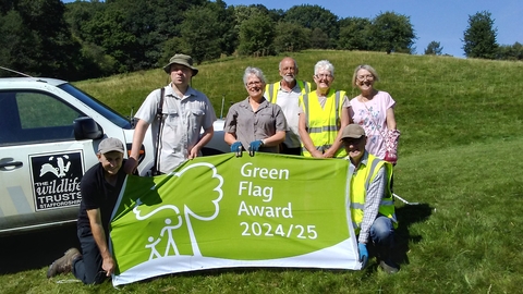 A group of people stand and crouch holding a large green and white flag which has the text "Green Flag Award 2024/25" on it. There's a grassy sloping hill behind them lined with trees and a white vehicle behind them with a black and white Wildlife Trusts logo on the side.