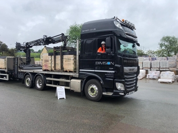 A black HGV lorry with numerous loads of brick stacks parked in a yard with a smiling happy driver who has long dark hair and wears a white hard hat