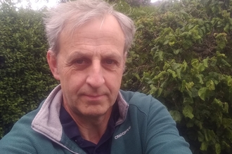 A man with grey hair in a green zip neck top stands in front of a green hedge