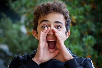 A teenage boy with curly dark brown hair shouts with his hands cupping his mouth to amplify his voice. Greenery behind him.