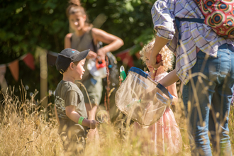 Two young children in a meadow with long grass hunting for bugs with a net and a woman holding it