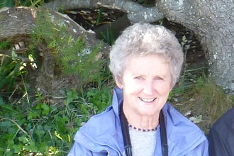 A woman smiles at the camera as she sits beneath a tree, she wears binoculars and a blue/lilac coat and has short grey hair.