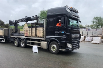 A black HGV lorry with numerous loads of brick stacks parked in a yard with a smiling happy driver who has long dark hair and wears a white hard hat