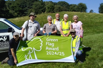 A group of people stand and crouch holding a large green and white flag which has the text "Green Flag Award 2024/25" on it. There's a grassy sloping hill behind them lined with trees and a white vehicle behind them with a black and white Wildlife Trusts logo on the side.