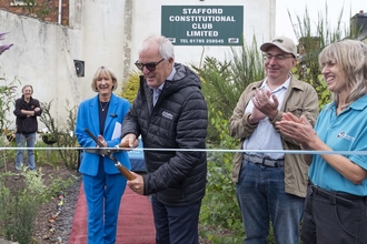 A group of people watch and clap as a man cuts a ribbon with some garden shears. They stand on a red carpet in a community garden and are all smiling.