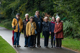 A group of people stand on a paved pathway smiling at the camera. They are mostly elderly, with one younger man stood in the middle of the group who wears a black SWT branded waterproof jacket.