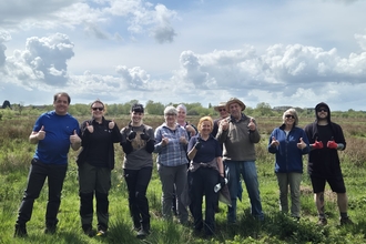 A large group of people smiling at the camera giving thumbs up! They stand in an outdoor area with a wetland behind them and cloudy sky overhead