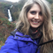 A woman with long blonde hair smiles at the viewer as she stands in front of a waterfall cascading down a hillside. She wears a bright blue coat.