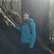 A woman stands with her hand resting on a large tree in bright sunlight in a woodland. She wears a woollen hat and blue waterproof top.