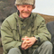 A man aged around 50/60 sits on a rock in an outdoor setting. He wears a green waterproof coat and a fur lined hat and is smiling at the camera.