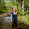 A woman with long hair tied back stands in a brook on a sunny early summer day. She has on dark trousers and welly boots. Behind her vibrant green trees extend along a dirt path into the background