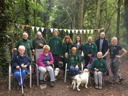 A group of woman and men stand and sit in two rows in a woodland setting with bunting strung up between the trees