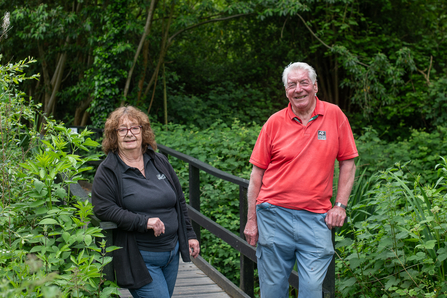A woman and man stand leaning on railings of a bridge surrounded by greenery. The woman wears a black polo, long sleeved jacket and jeans, she has dark curly hair and wears glasses. The man has grey hair and wears a red polo top and grey/blue trousers.