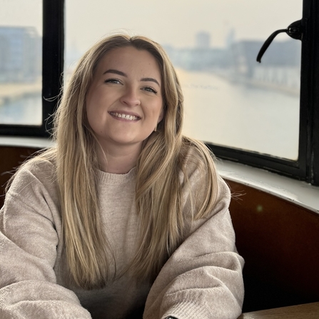 A woman sits by a window overlooking a river/water. She wears a cream/beige jumper and has long blonde hair. She smiles at the camera.