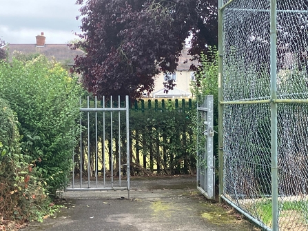 A suburban gate opens up onto a path near former tennis courts.