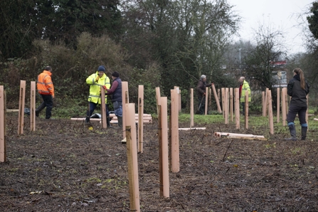 A group of people in outdoor wear are out in a muddy field surrounded by trees and tree guards.