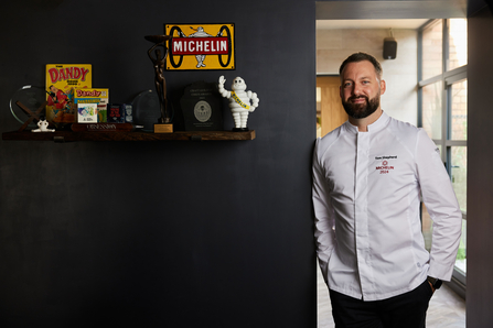 A man stands smiling at the viewer as he leans against a black painted wall which has a shelf hosting various ornaments including a white michelin man figure. He wears a white chefs top.