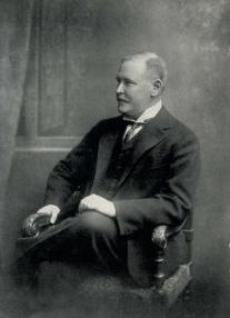 A black and white digital scan of an analogue portrait of a wealthy man in a suit looking to the left as he sits in a chair.