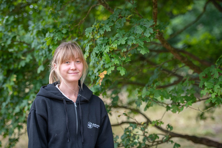 A woman with blonde hair tied in a pony tail stands in front of a tree smiling. She wears a black hooded top with Staffordshire Wildlife Trust's logo on it.