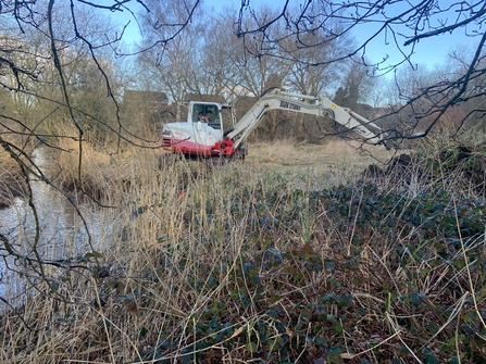 A man operates a piece of machinery near a brook in an area filled with vegetation.