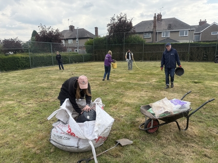 A group of people collect seed from a large tonne bag and distribute over a disused tennis court.