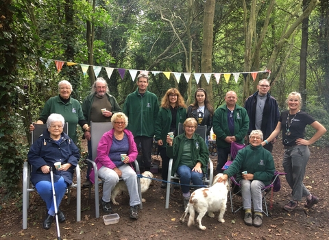 A group of woman and men stand and sit in two rows in a woodland setting with bunting strung up between the trees