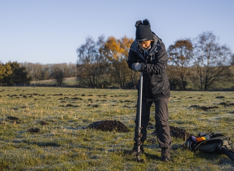 A woman dressed in dark clothing and a woolly hat pushes a long device into the earth in a big open space with trees in the background