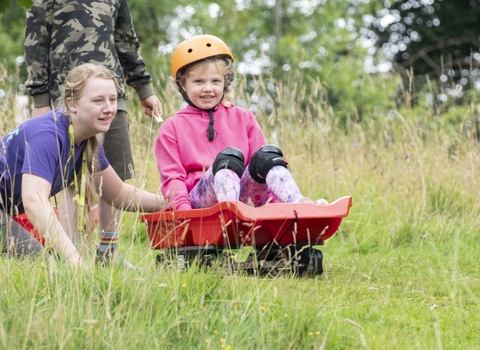 A girl sits on a red grass sled and looks excited as she rolls down a hill, a young woman in purple pushes her