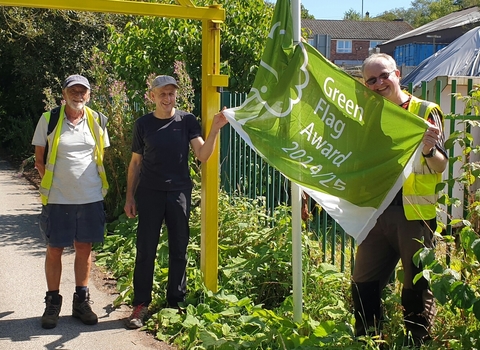 Three people stand by a yellow car park barrier and flag pole as they raise a green and white flag, one of the people - a man with sunglasses and a high vis tabbard holds the flag up and smiles.