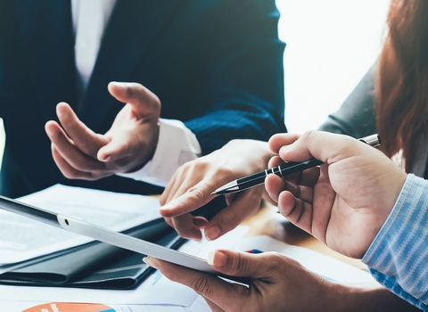 Three people sit in close proximity with documents and a tablet in hand, along with a pen. They're in formal wear suggesting a business meeting.