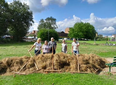 A group of 5 people standing with rakes and large amounts of hay piled up as high as their waists. They stand in a green field with trees in the distance and houses nearby.